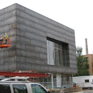 Modern concrete building under construction; orange lift with workers, large window, cloudy sky.