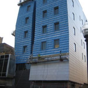 Building under construction; blue and silver facade with windows. Workers on a suspended platform.