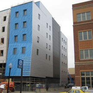 Building under construction with blue, silver, and dark gray panels, scaffolding, and adjacent buildings.