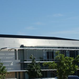 Modern building with a flat roof, reflective metallic siding, large windows, and trees against a blue sky.