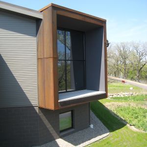 Modern house exterior with large, brown-framed window box. Gray siding, green lawn, trees in background.