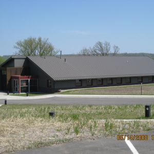 Building with dark metal roof and entry canopy, on a grassy hillside, blue sky background.