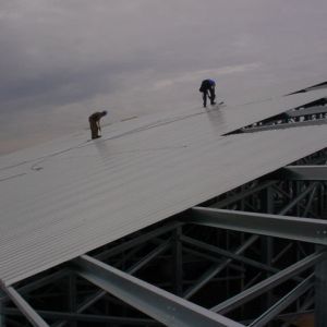 Two workers on a metal roof, installing panels. Steel framework visible against a cloudy sky.