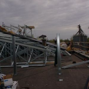 Metal framework construction on a rooftop with workers and silos visible under a cloudy sky.