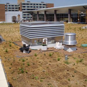 Rooftop equipment on a green roof, with building in the background. Gray and silver ventilation units, brown grass.