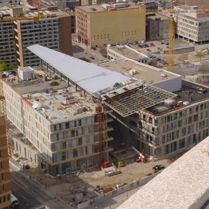Building under construction, three stories high with a long, slanted roof, surrounded by other buildings and construction equipment.
