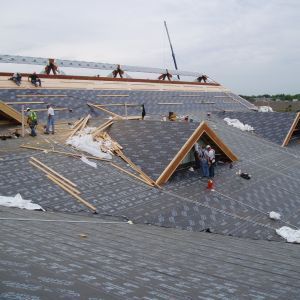 Roofers installing shingles on a large building. Several workers on the roof, with construction materials and tools visible.