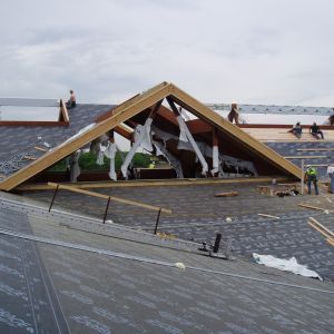 Roof construction: workers on a roof, wooden frame of a dormer, gray shingles, cloudy sky.