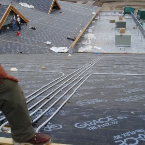 Construction workers on a roof, installing pipes over black underlayment. Another roof and equipment are visible.