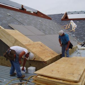 Two construction workers building on a roof, installing plywood. Gray roof, overcast sky.