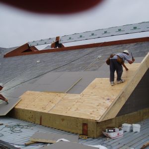 Construction workers on a roof, installing wooden framework. Gray roofing, cloudy sky.