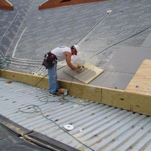 Construction worker on a corrugated metal roof, using a drill. He's wearing a hard hat, working with wood.