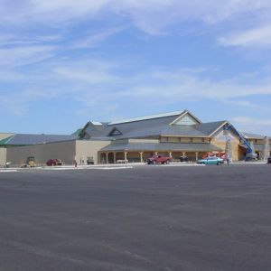 Large beige building with dark roof, open porch, and empty asphalt parking lot under blue sky.
