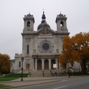 Large stone cathedral with two bell towers, a dome, and steps leading to the entrance. Fall trees frame the building.