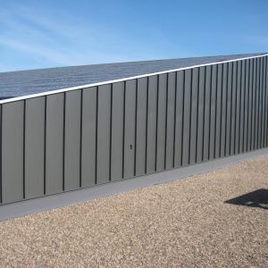 Gray metal-clad building with a slanted roof covered in solar panels, set against a blue sky.