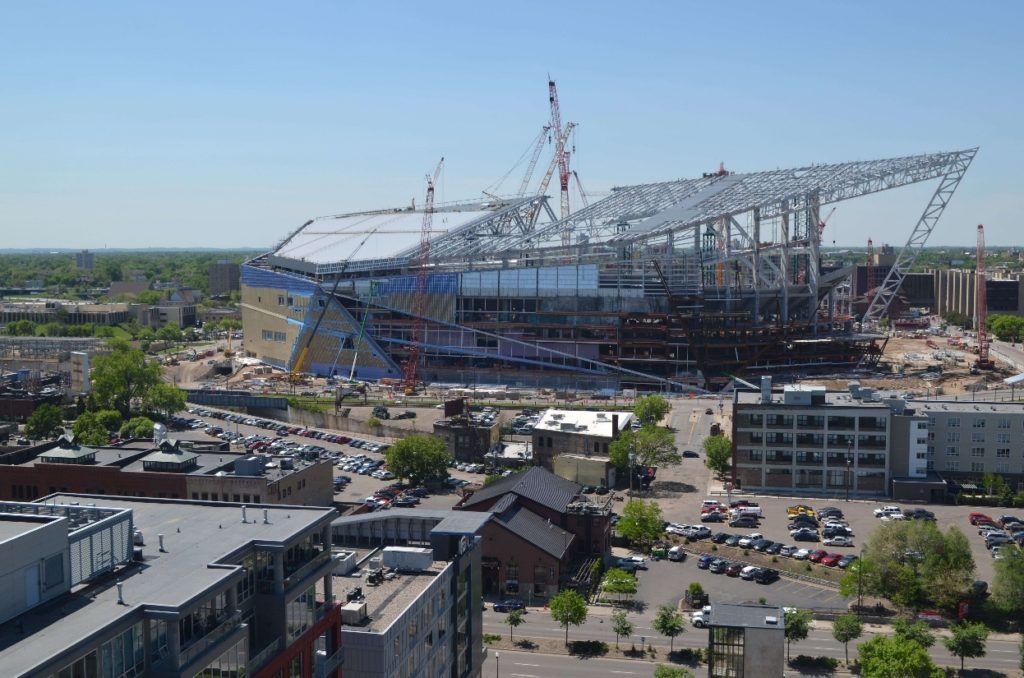 Stadium under construction, with cranes, metal framework, and surrounding buildings. Blue sky overhead.