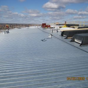 Construction site roof covered in corrugated metal. Workers and equipment visible under a cloudy sky.