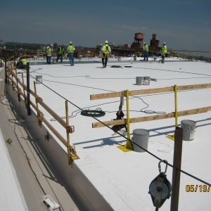 Construction workers on a white rooftop, wearing safety gear and near temporary guardrails and cables.