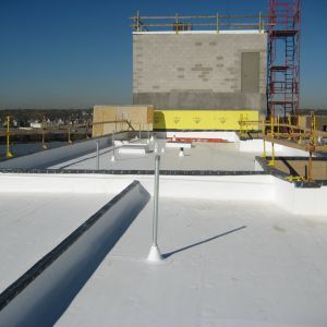 White flat roof with vents and guardrails, construction site in the background. Sunny day.
