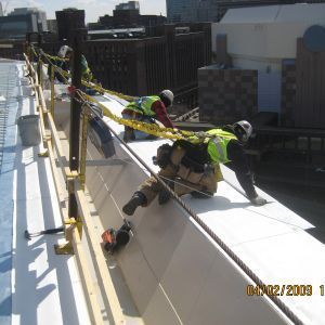 Construction workers on a roof, attached to safety lines, working on a white surface next to a safety railing.