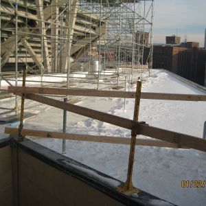 Snowy rooftop with wooden safety rails, scaffolding, and a city skyline in the background.