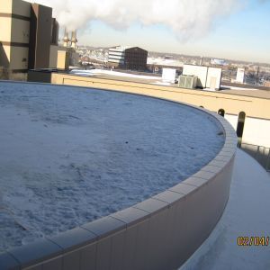 A rooftop with snow covering it, a curved wall, and city buildings in the background.