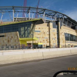 Exterior of a stadium under construction with tan stone facade, glass windows, and yellow accents.