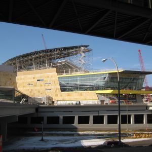 Construction site with tan building and glass sections, under blue sky; crane visible.