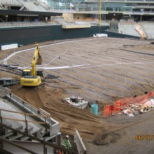 Construction of a baseball field. An excavator on dirt, lines drawn for the field, stadium in the background.