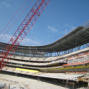 Construction of a large stadium. Red crane extends over the concrete and steel structure against a blue sky.
