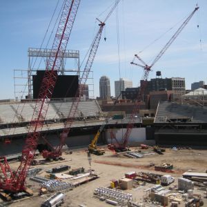 Construction site with multiple red cranes, building a stadium. Buildings and blue sky in the background.