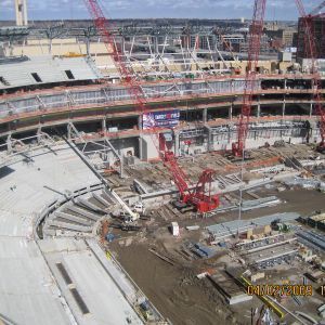 Construction site of a large stadium; cranes lifting materials, various stages of building visible.