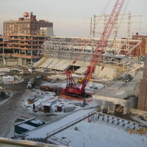 Construction site with red crane lifting materials, stadium structure under construction; snow on ground.