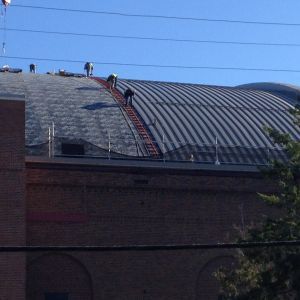 Workers on a curved metal roof, climbing a ladder. Brick building below, blue sky overhead.