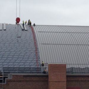 Construction workers on curved metal roof, using crane to lift materials, overcast sky.