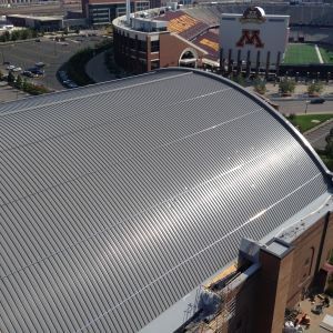 Curved, metallic roof of a stadium with a football stadium in the background. Sunny day.