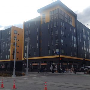 Modern, multi-story building with black and yellow exterior, located on a street corner. Pedestrians and traffic visible.