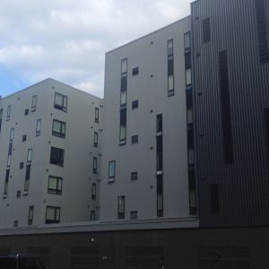 Modern apartment buildings with rectangular and vertical windows, gray exteriors, and a cloudy sky.