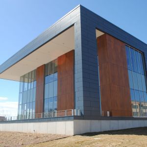 Modern building with glass walls, weathered metal panels, and dark gray roof against a clear blue sky.