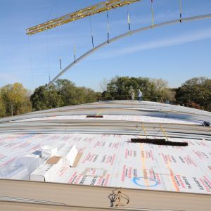 Construction workers installing curved metal framework on a domed roof covered with white insulation panels.