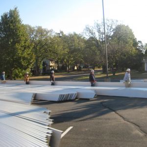 Construction workers handling large white panels outdoors on a road, with trees and houses in the background.