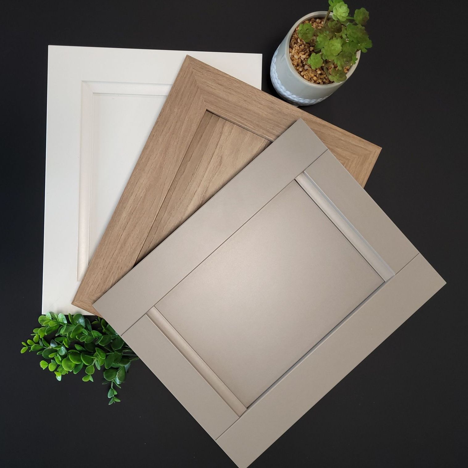 Trois échantillons de portes d'armoires en blanc, en similibois et en gris, sur une surface noire avec de la verdure.