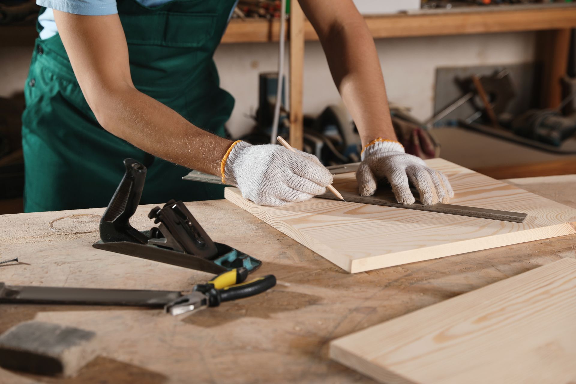 Personne portant un tablier vert et des gants utilisant une règle pour mesurer et marquer le bois sur un établi.