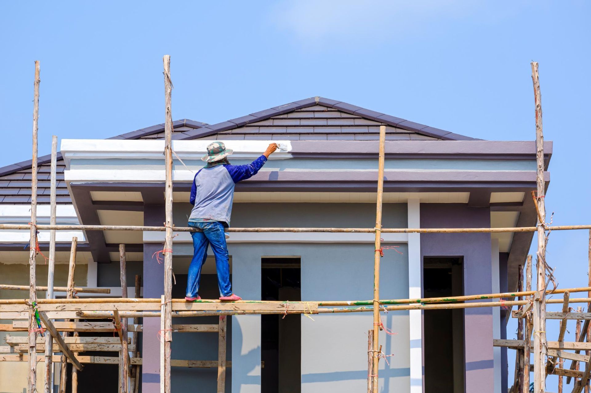 Construction worker painting trim on a house, standing on scaffolding, clear blue sky.