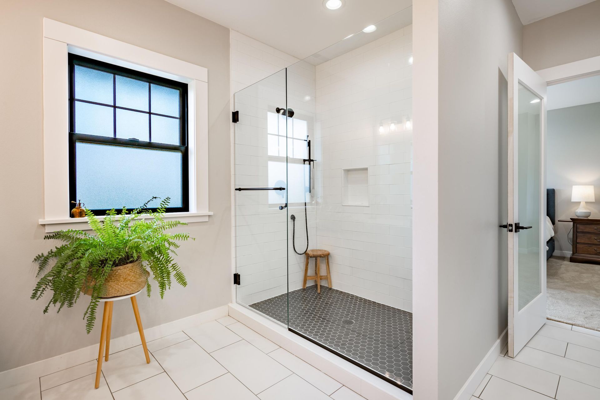 Bathroom with glass shower, white tile, black window frame, and fern in a basket on a wooden stand.