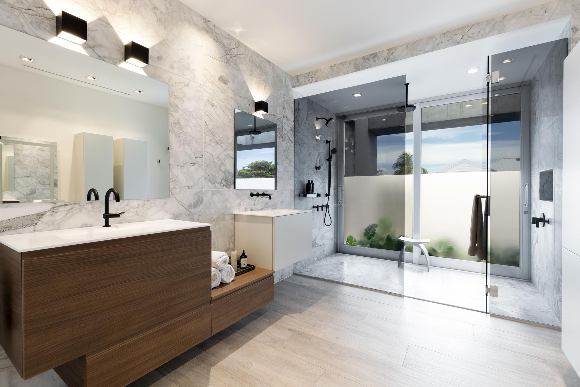 Modern bathroom with marble walls, wooden vanity, glass shower, and outdoor view.