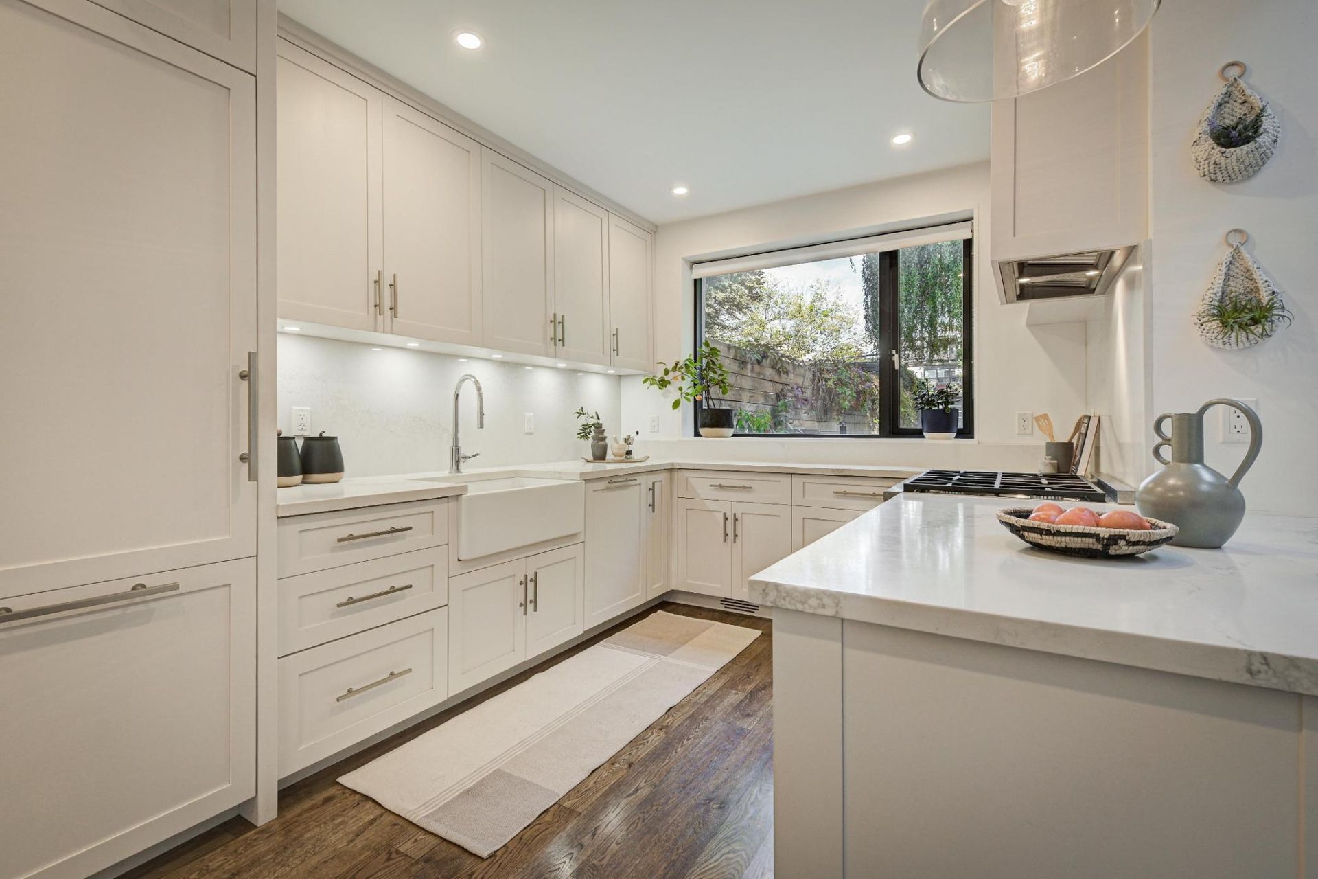 White kitchen with marble countertops, wood floors, and a window overlooking greenery.