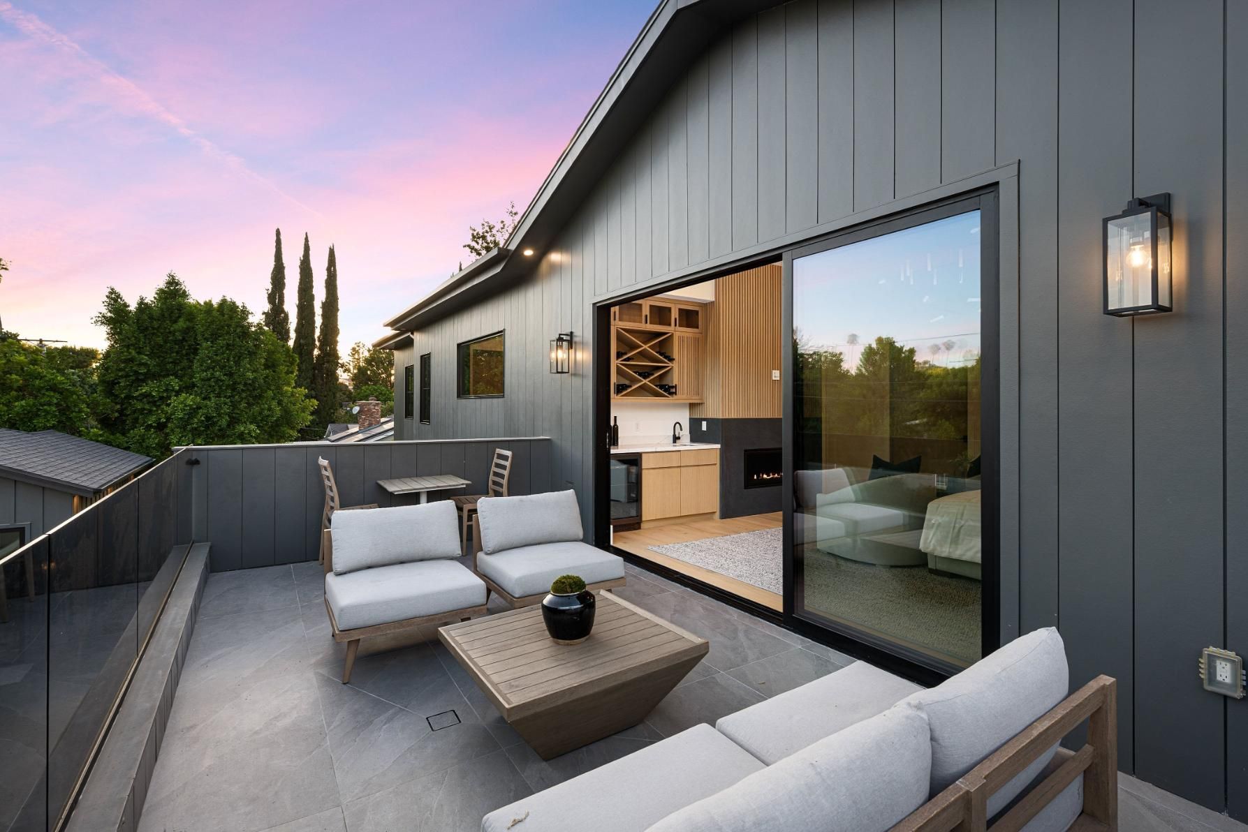 Rooftop patio with seating, glass railing, and access to a modern interior, against a twilight sky.