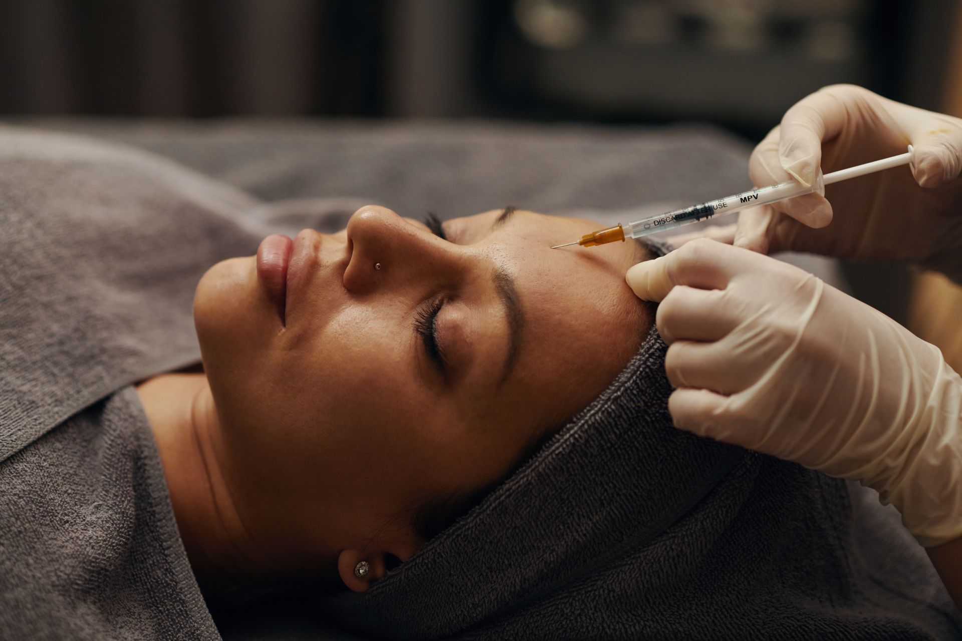 A person in a medical setting receives a cosmetic injection on their forehead from a practitioner wearing gloves.