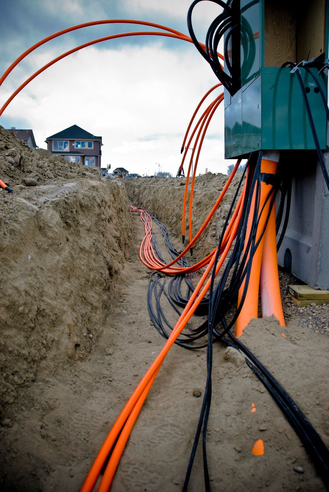 Trench with orange conduits and black cables; construction site, house in background.